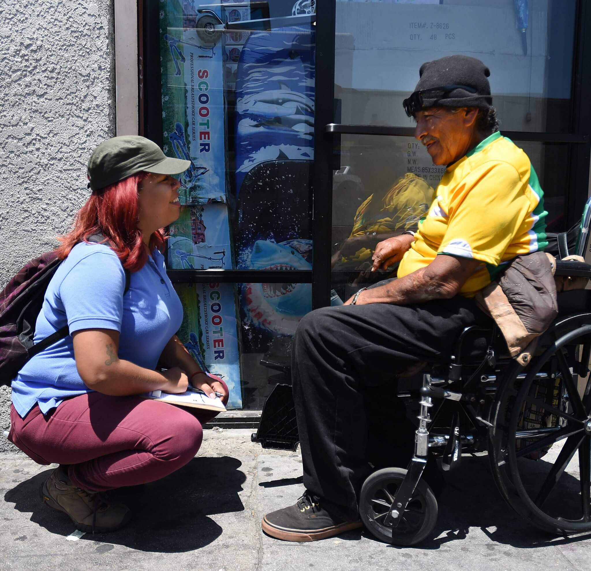 A young woman squats in front of an older man in a wheelchair as they smile at each other.